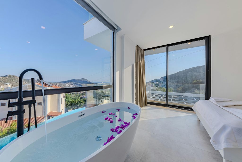 Modern hotel bathroom with freestanding tub filled with petals, floor-to-ceiling windows, and mountain views